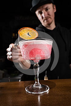 Bartender is decorating pink Clover club alcoholic cocktail with orange slice at the bar. Bartender mixes egg white