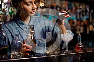Bartender girl pouring to the measuring glass cup with ice cubes a brown alcoholic drink from steel jigger