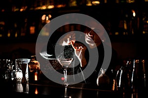 Bartender girl sprinkling on a alcoholic drink in the cocktail glass with a orange zest juice