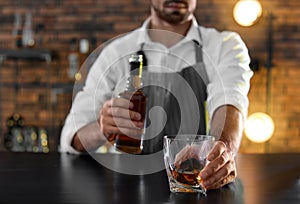 Bartender with glass and bottle of whiskey at counter in bar, closeup.