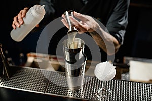 Bartender pouring a alcoholic drink from the jigger to a steel shaker holding a white plastic bottle