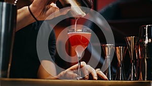 Bartender pours an ingredient into a red cocktail on the bar counter