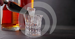 bartender puts cold ice cubes for cooling into a glass whiskey glass on a background of bottles