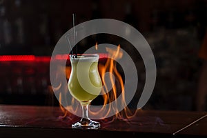 Bartender sprinkles on illuminated glass with bright green cold cocktail on bar counter and makes fire flame over it