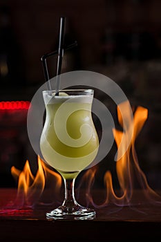 Bartender sprinkles on illuminated glass with bright green cold cocktail on bar counter and makes fire flame over it
