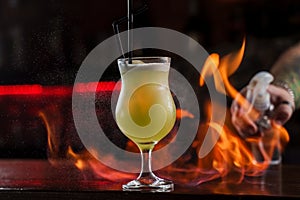 Bartender sprinkles on illuminated glass with bright green cold cocktail on bar counter and makes fire flame over it