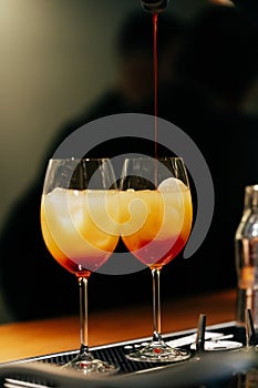 On the bartenders table are two glasses of orange and red cocktails into which the drink is poured. Orange juice, alcohol. On a