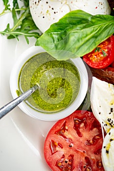 Basil pesto sauce in a small bowl, with fresh basil leaves, tomato and mozzarella cheese