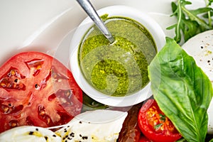 Basil pesto sauce in a small bowl, with fresh basil leaves, tomato and mozzarella cheese