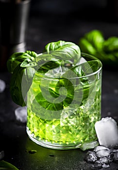 Basil smash green alcoholic cocktail drink with dry gin, sugar syrup, lemon, basil and ice, dark bar counter background