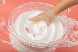 A Basin with Soapy Water, Hand Holds Soapy Sponge with Foam on Pink Background