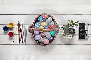 Basket with colorful Easter eggs, painted in handmade, bouquet of spring flowers, paints, brushes and retro camera stand in a row
