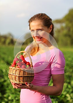 Basket full of red ripe strawberries, blurred young woman holding it, and strawberry fields in background.