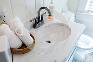 A basket of organized clean rolled white towels on a bathroom counter in a guest bathroom near a sink and toilet.
