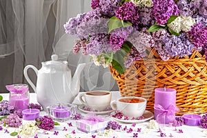 A basket of purple flowers sits on a table with a white tea set and candles