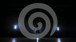 The basketball player stands on a dark playground and holds the ball in his hands and looks into the camera in the dark