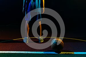 Basketball. A teenage boy in a blue sportswear stands on the Playground with ball. Close up of legs. Black background. Rear view.