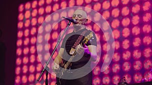 Bass guitarist sings on stage into microphone at pop band performance. Stage is illuminated by blue pink and red lights