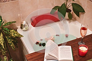 Bath with flower petals. Book, candles and glass of wine on a wood tray. Organic Spa Relaxation in comfort cozy bathroom
