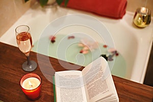 Bath with flower petals. Book, candles and glass of wine on a wood tray. Organic Spa Relaxation in comfort cozy bathroom