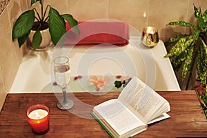 Bath with flower petals. Book, candles and glass of wine on a wood tray. Organic Spa Relaxation in comfort cozy bathroom