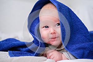 Bath time. Cute smiling caucasian infant baby looking at camera in blue towel after bathing in the bath on white bed