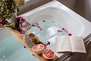 Bath tub with a tray with grapefruit slices, bunch of grapes, a glass of wine and a book