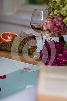 Bath tub with a tray with grapefruit slices, bunch of grapes, a glass of wine and a book, selective focus