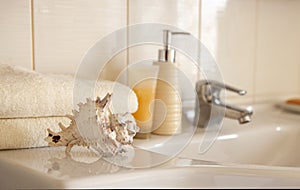 Bath white cotton towels, ceramic bottle with shampoo, tube with cream and cockleshell on Blurred bathroom interior background