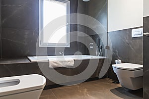 Bathroom with brown marble, window with natural light, white bathtub, toilet and bidet.