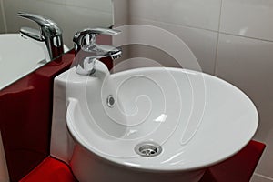 Bathroom interior with round sink and faucet on red stand and white tile background.