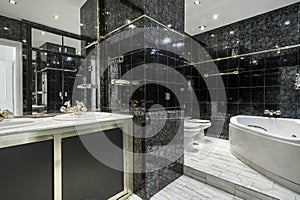 Bathroom with white porcelain sink above chest of drawers and beveled mirror above wall sconce in a room with black tiles and a