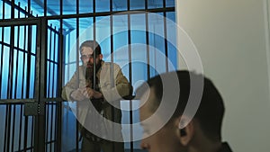 A battered homeless man with a beard stands behind a prison grate and looks at the policeman.