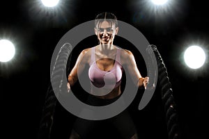 Battle ropes. Fit sports woman, athlete working out in functional training doing exercise on black background. Fitness