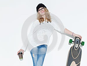 Bautiful fashionable cheerful young girl in a white sweater with a glass of coffee and longboard posing near white wall. In the st