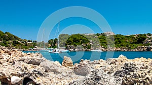 A bay in the Mediterranean with sailing ships and rocks in the foreground