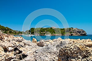 A bay in the Mediterranean with sailing ships and rocks in the foreground