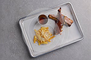 BBQ marinated spareribs with hot spice sauce and french fries served on a gray metal tray over grey textured background.