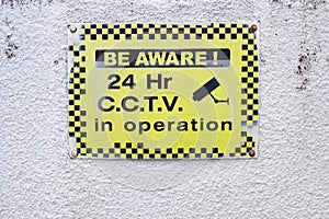 Be aware 24 hr hour CCTV security camera in operation yellow sign on white wall