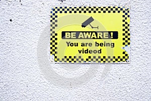 Be aware 24 hr hour CCTV security camera in operation yellow sign on white wall