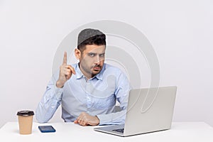 Be careful! Serious bossy businessman sitting office workplace with laptop on desk, showing finger up warn gesture