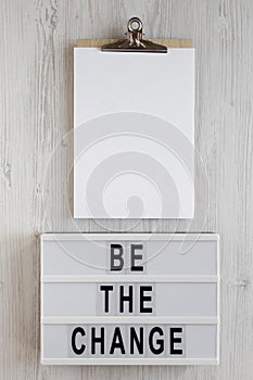 `Be the change` words on a lightbox, clipboard with blank sheet of paper on a white wooden surface, top view. Overhead, from abo