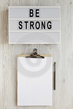 `Be strong` words on a lightbox, noticepad with blank sheet of paper on a white wooden background, top view. Overhead, from abov