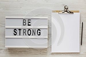 `Be strong` words on a lightbox, noticepad with blank sheet of paper on a white wooden background, top view. Overhead, from abov