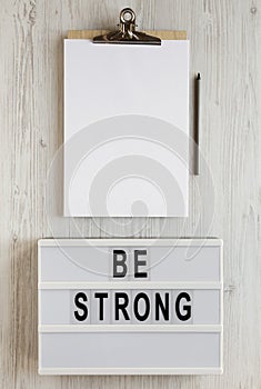 `Be strong` words on a modern board, noticepad with blank sheet of paper on a white wooden background, top view. Overhead, from