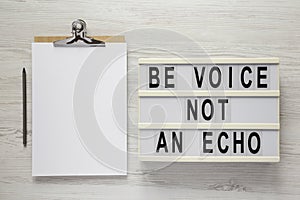 `Be voice not an echo` on a lightbox, clipboard with blank sheet of paper on a white wooden background, top view. Flat lay,