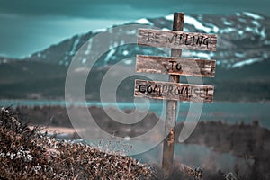 be willing to compromise text quote engraved on wooden signpost outdoors in landscape looking polluted