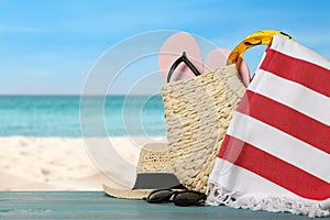 Beach bag with towel, flip flops, hat and sunglasses on light blue wooden surface near seashore. Space for text