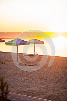 Beach chaise lounges under the umbrella on the sandy shore of the lake at sunset