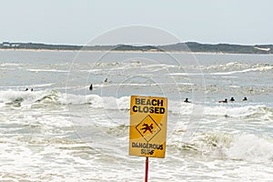 Beach closed sign. Despite the warning people are still swimming and surfing. Selective focus on